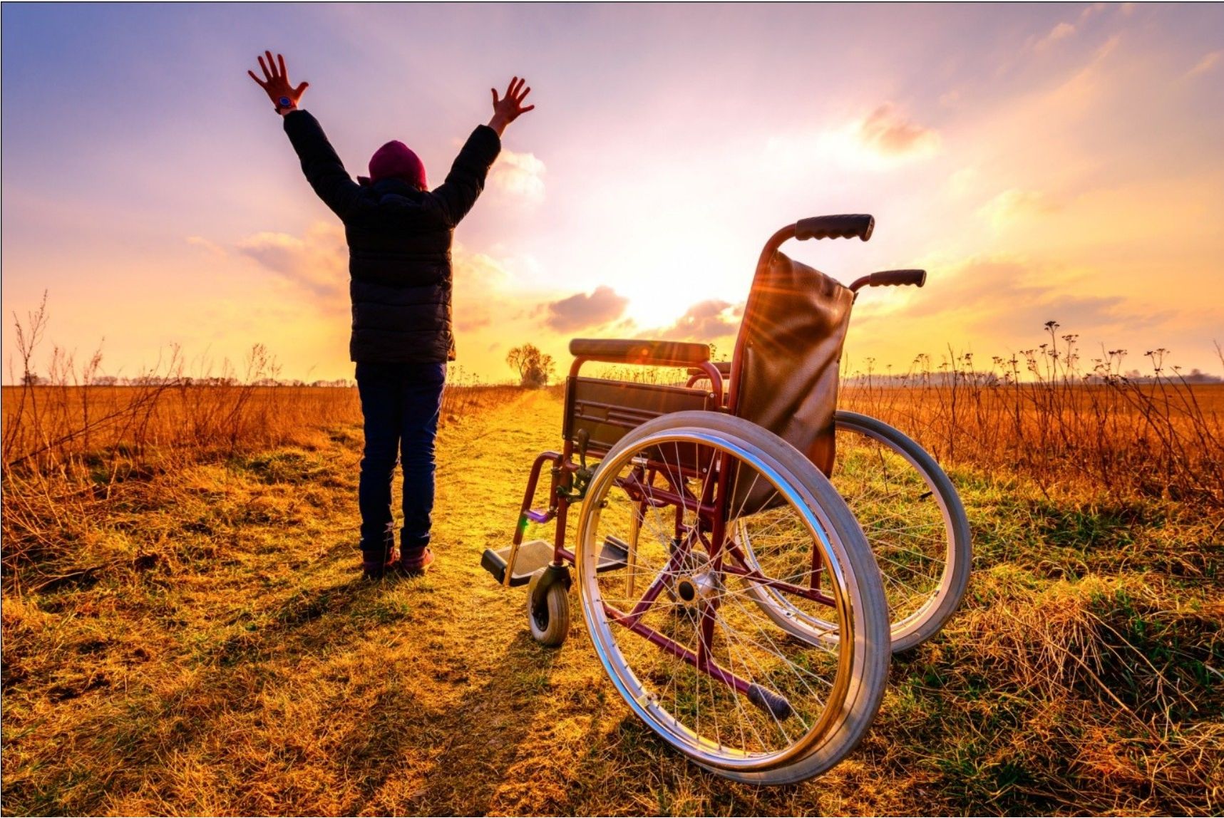 A person standing with arms raised in a field next to an empty wheelchair at sunset