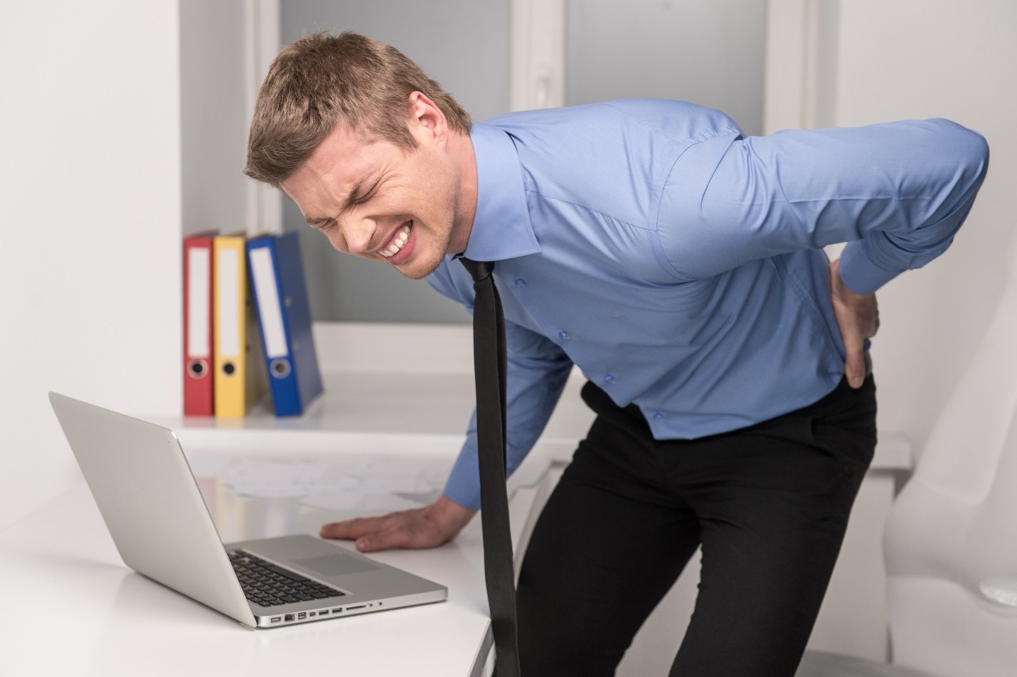 A man in a blue shirt and tie holding his lower back in pain while standing at a desk with a laptop