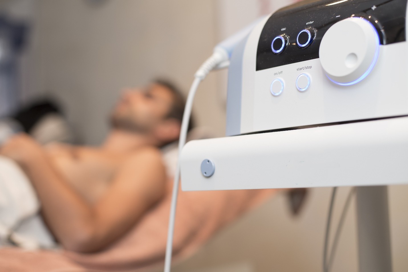 A medical device with on/off and start/stop buttons in the foreground, with a patient on a treatment table in the background
