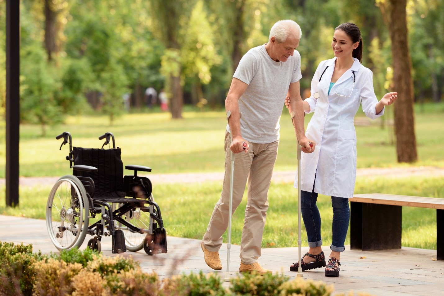 A doctor assisting an elderly man walking with crutches in a park, with a wheelchair nearby